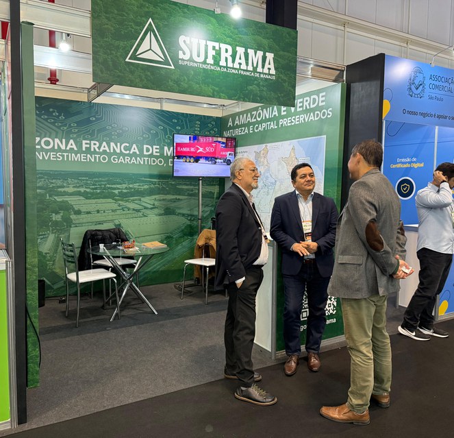 Foto mostra três homens conversando em frente um estande verde e branco