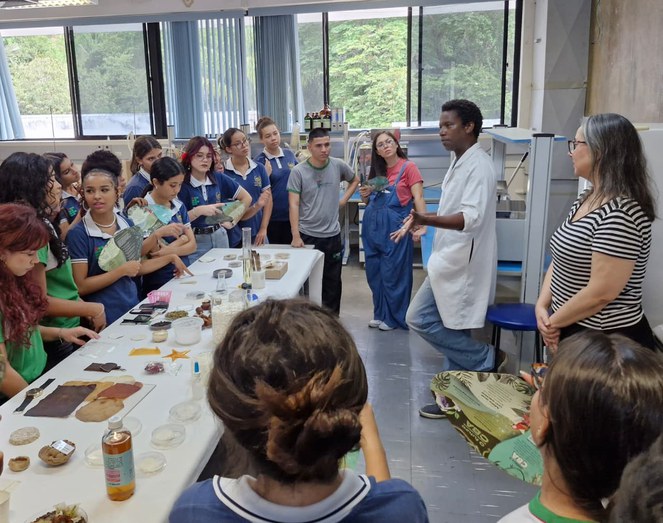 Foto mostra homem de jaleco branco fazendo uma explicação a grupo de estudantes em um laboratório