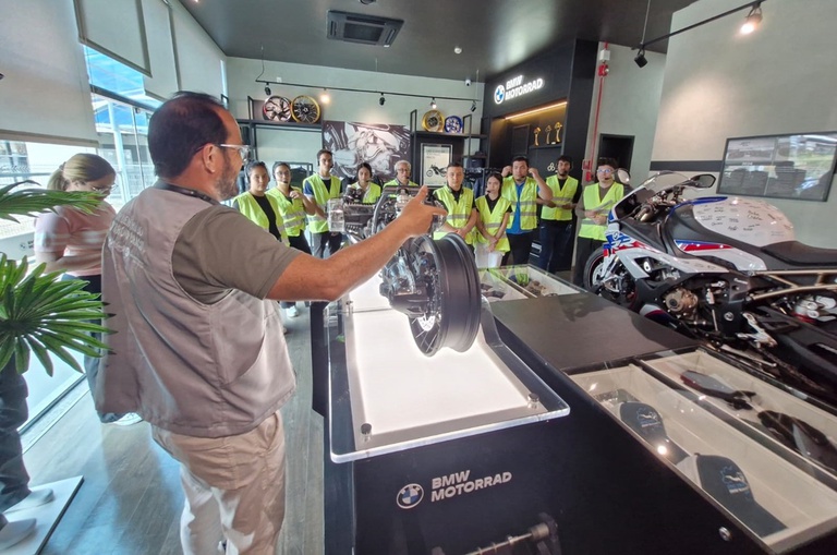Foto mostra homem falando jovens de colete verde sobre peça de uma motocicleta