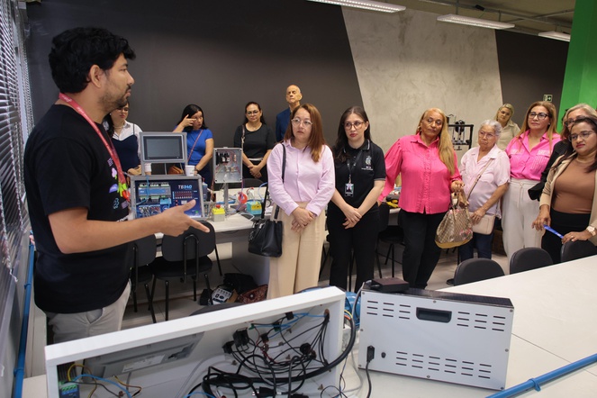 Foto mostra homem de blusa preta fazendo uma explanação a grupo de pessoas em meio a computadores