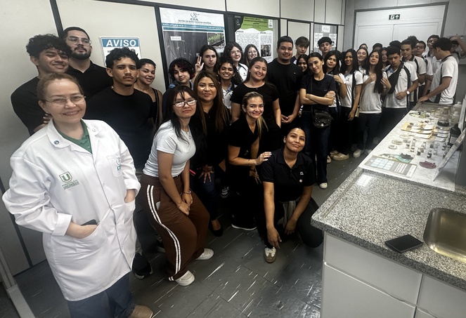 Foto mostra pesquisadora de jaleco branco posando ao lado de estudantes num laboratório de pesquisa