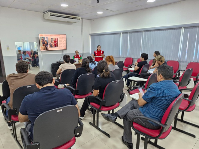 Foto mostra uma mulher em pé fazendo uma explanação a grupo de pessoas