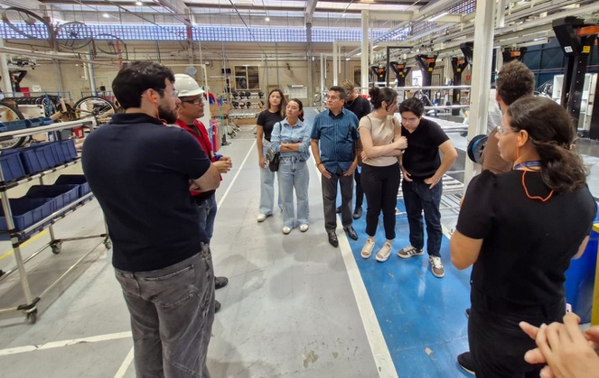 Foto mostra homem de jaqueta vermelha e capacete branco falando a um grupo de pessoas