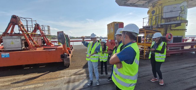 Foto mostra grupo de pessoas com jaquetas verdes e bonés em um porto flutuante