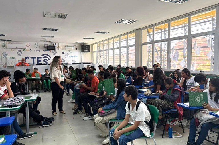 Foto mostra mulher em pé ministrando palestra para alunos