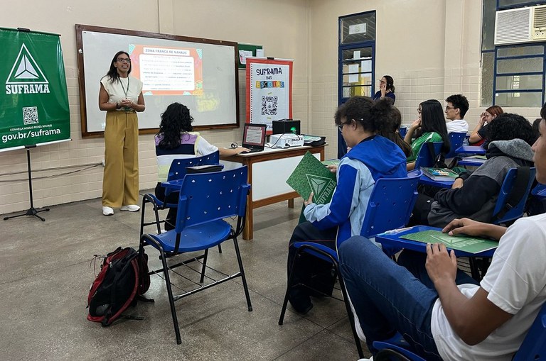 Foto mostra mulher de calça marrom e blusa bege ministrando palestra a alunos