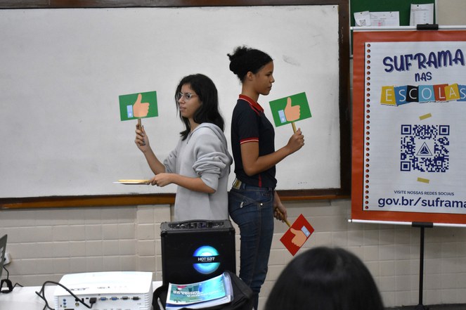 Foto mostra mulher de calça marrom e blusa bege ministrando palestra a alunos da rede estadual de ensino