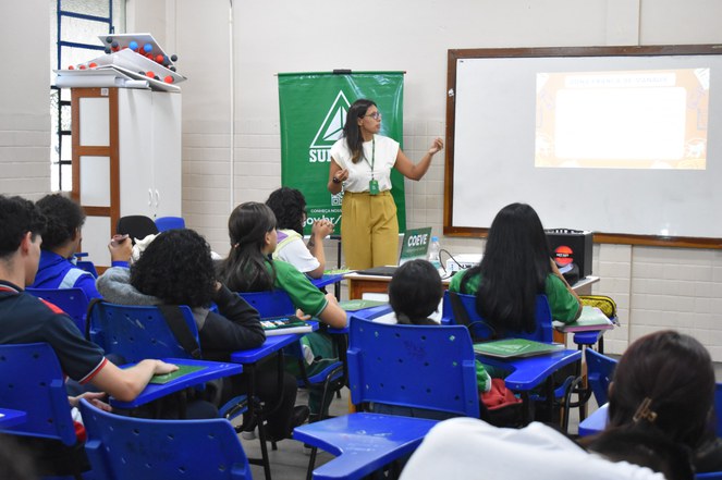 Foto mostra uma mulher de calça marrom e blusa bege ministrando palestra a alunos