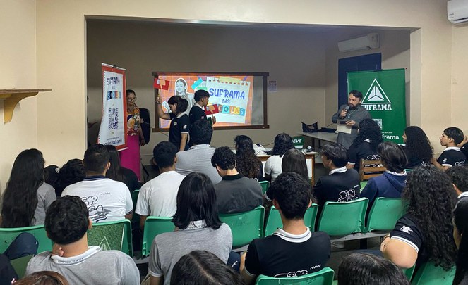 Foto mostra homem com microfone na mão e dois adolescentes segurando dois objetos, com um telão ao fundo