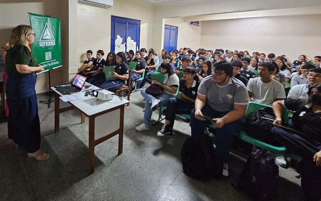 Foto mostra mulher loira, vestida com blusa verde e saia azul, e observada por um grupo de estudantes