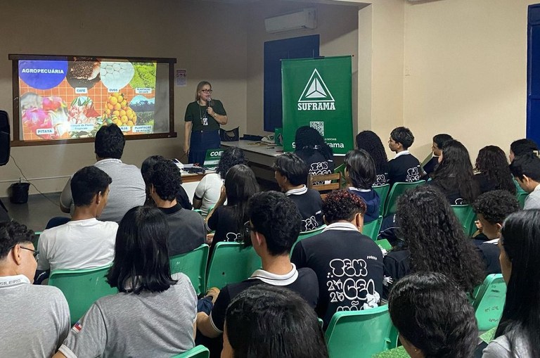 Foto mostra mulher com microfone na mão, um telão de fundo e adolescentes assistindo a explicação dela