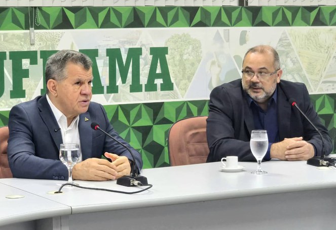 Foto mostra homem de blazer falando ao microfone observado por outro homem