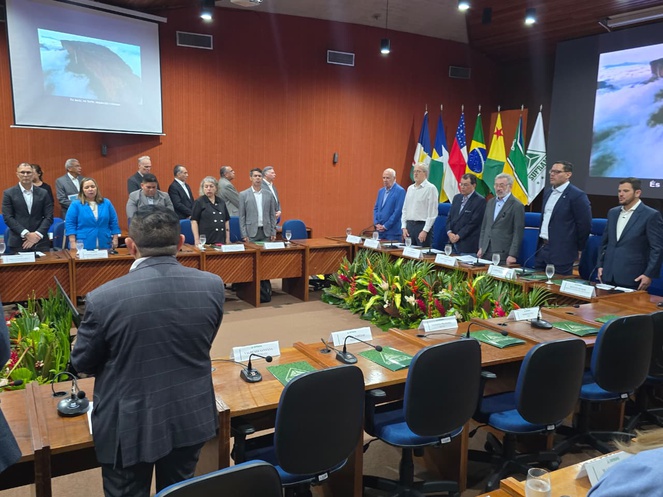 Foto mostra pessoas em pé cantando o Hino Nacional Brasileiro durante evento em um auditório