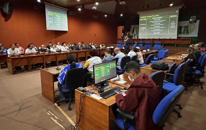 Foto mostra homem em pé falando ao microfone num auditório e com um painel grande ao fundo e pessoas ao redor