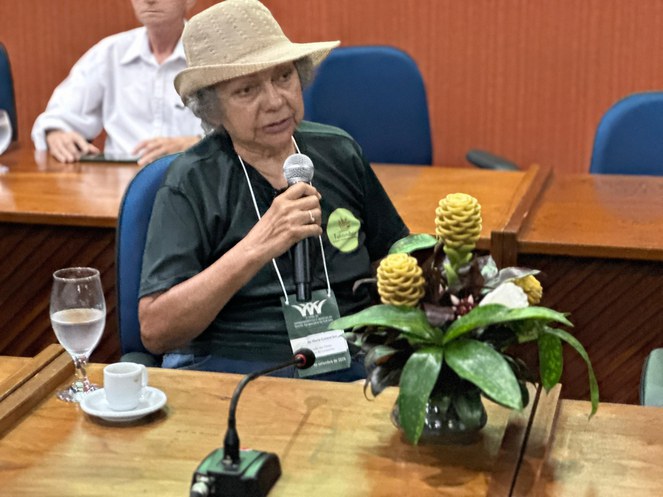 Foto mostra uma mulher falando ao microfone com uma planta na frente dela