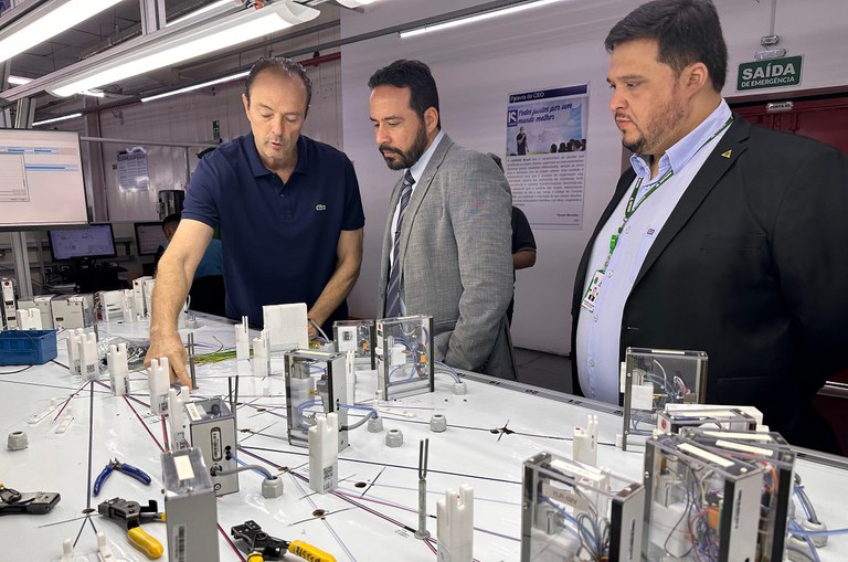 Foto mostra homem de blusa azul escuro mostrando equipamentos eétricos a dois homens vestidos com blazer