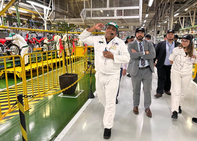 Foto mostra homem de blusa e calça brancas e microfone à boca explanando a visitantes dentro de uma fábrica