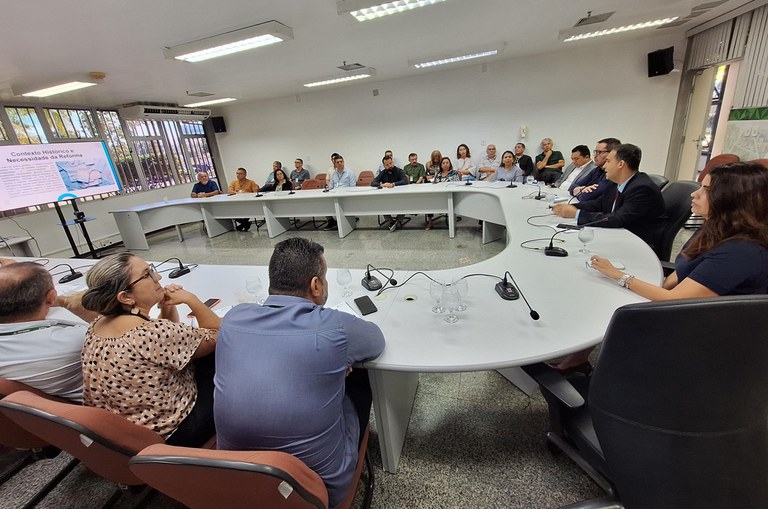 Foto mostra homem de paletó escuro falando ao microfone, observado por um grupo de pessoas em uma reunião presencial e online