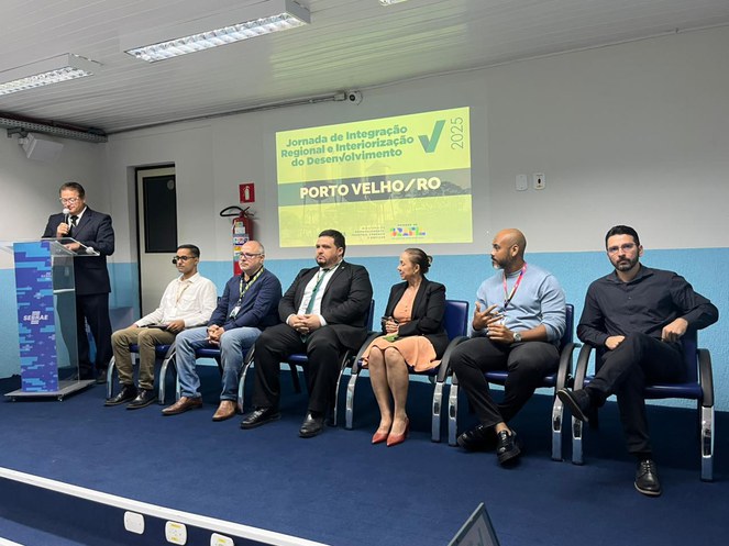 Foto mostra homem em pé falando ao microfone ao lado de um grupo de pessoas