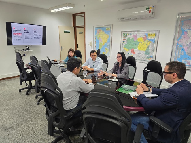 Foto mostra grupo de pessoas em reunião numa sala com um telão ao fundo como apoio