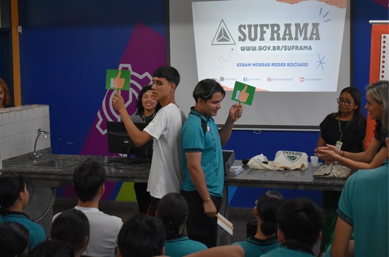 Foto mostra dois jovens segurando dois objetos, observados por outras pessoas