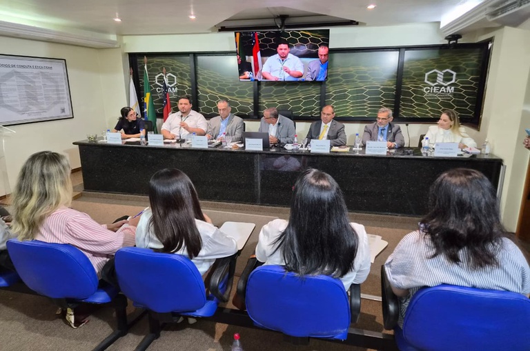 Foto mostra homem de camisa branca falando ao microfone para outra pessoas, numa sala com telão