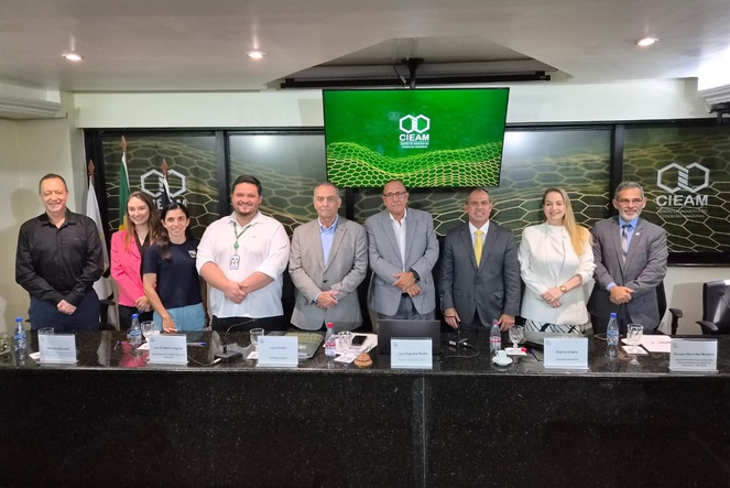 Foto mostra pessoas em pé, tendo como pano de fundo um painel e um telão