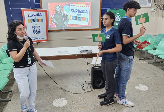 Foto mostra jovem de blusa preta e microfone na mão fazendo uma dinâmica com outros dois jovens uma escola