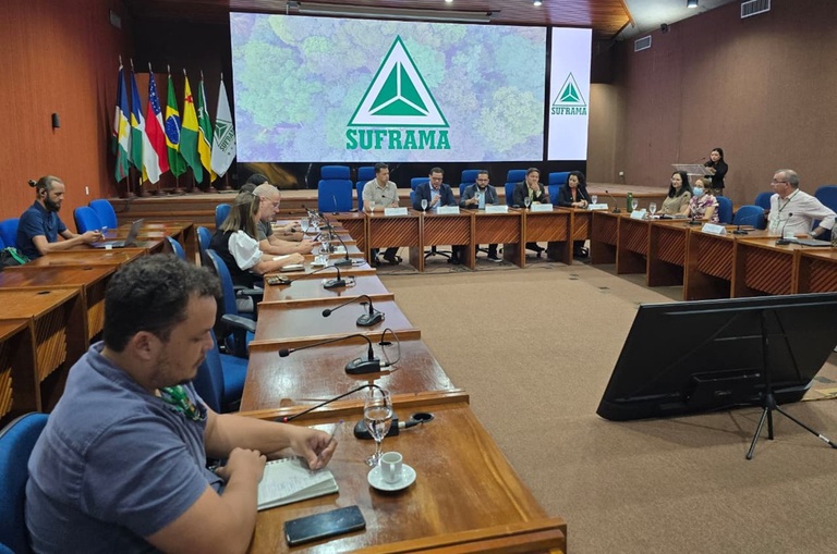 Foto mostra pessoas sentadas numa reunião, com um telão ao fundo