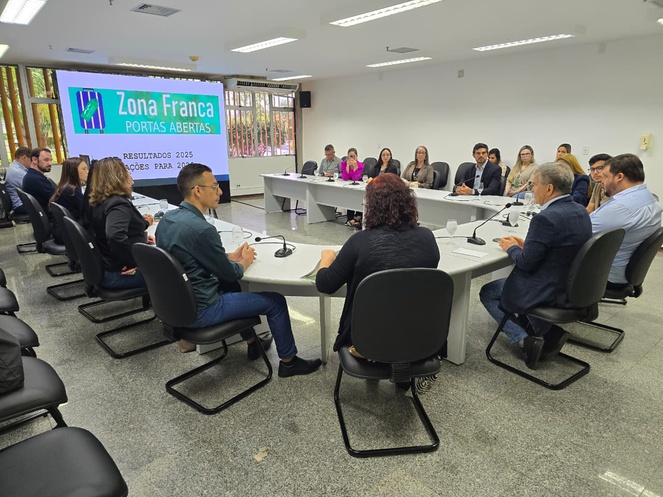 Foto mostra pessoas à mesa numa reunião com o apoio de um telão