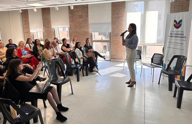 Foto mostra uma mulher em pé com microfone na mão falando a um grupo de outras mulheres sentadas