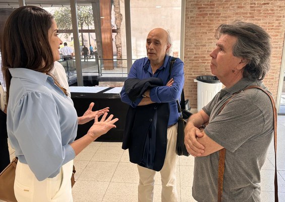 Foto mostra uma mulher em pé de blusa azul falando com dois homens