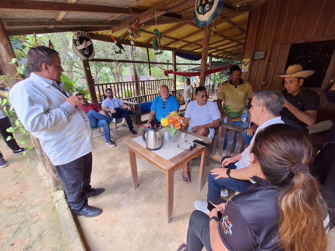 Foto mostra um homem de blusa branca e sentado à mesa (centro), conversando com grupo de pessoas