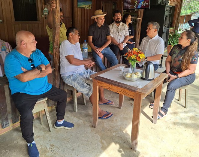 Foto mostra homem de blusa branca e sentado à mesa (centro), conversando com grupo de pessoas