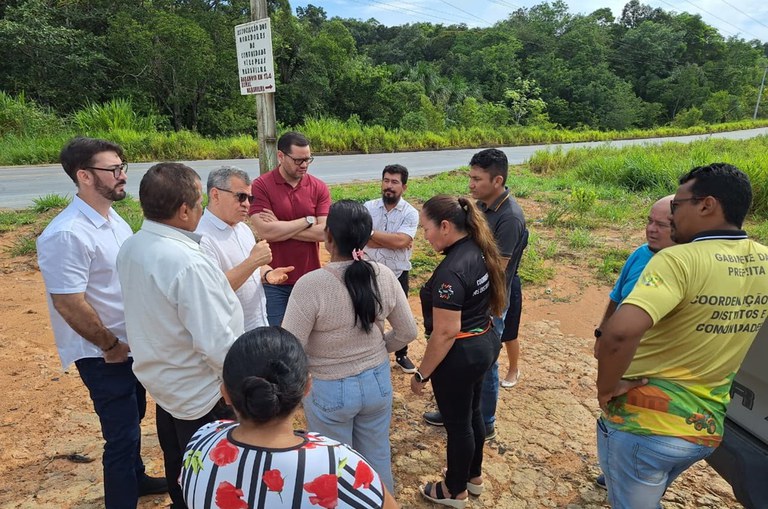 Foto mostra homem de blusa branca e óculos escuros conversando com grupo de pessoas, tendo uma placa sinalizadora ao fundo