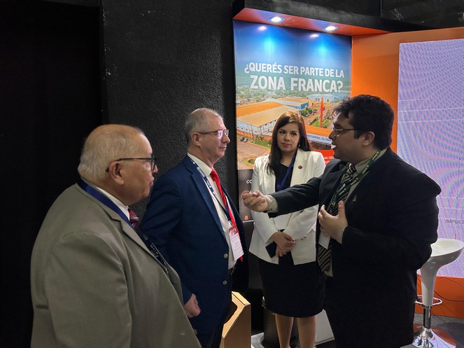 Foto mostra homem de blazer e óculos conversando com outros dois homens, observados por uma mulher