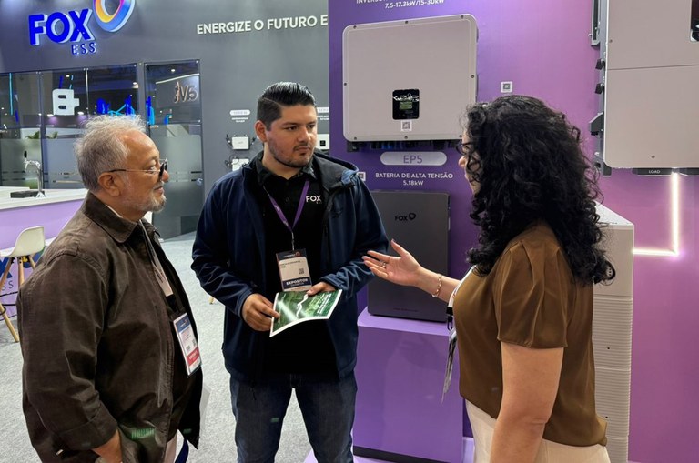 Foto mostra uma mulher dando uma explicação a um homem, observada  por outro homem