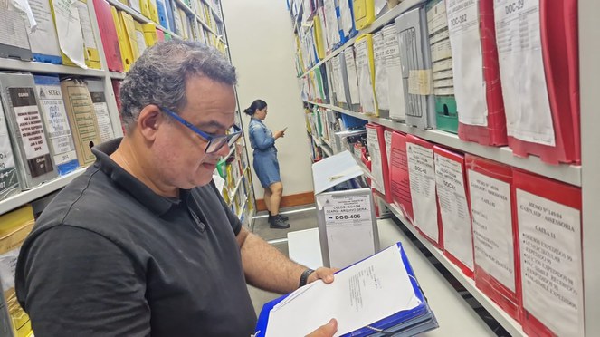 Foto mostra homem de óculos olhando para uma papelada, num ambiente cheio de caixas documentais