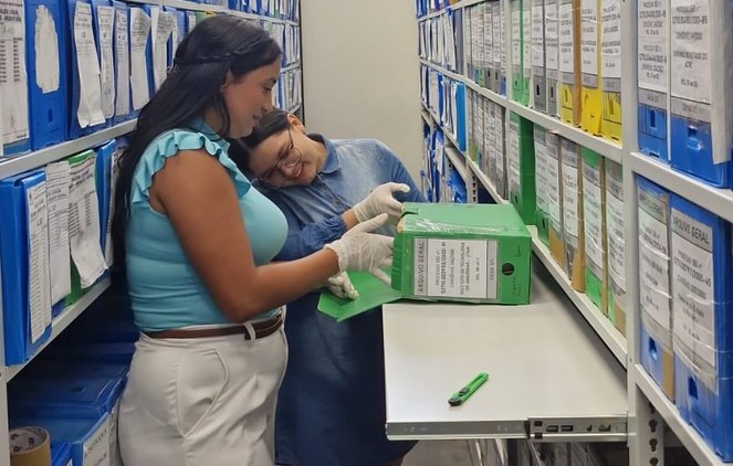 Foto mostra duas mulheres com luvas brancas, verificando uma caixa documental