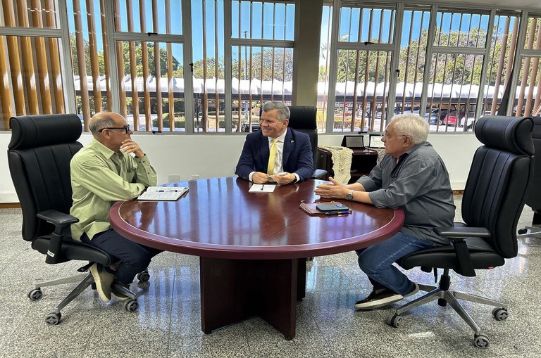 Três homens de meia idade reunidos sentados ao redor de uma mesa redonda
