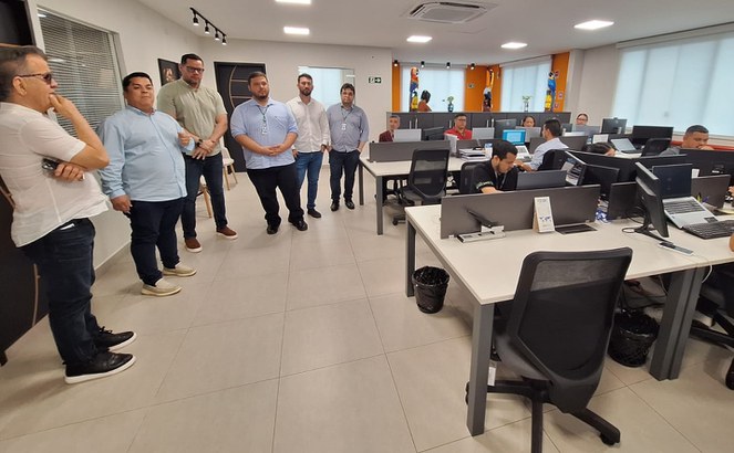 Foto mostra homem de óculos escuro com a mão no queixo e observando um grupo de pessoas numa sala