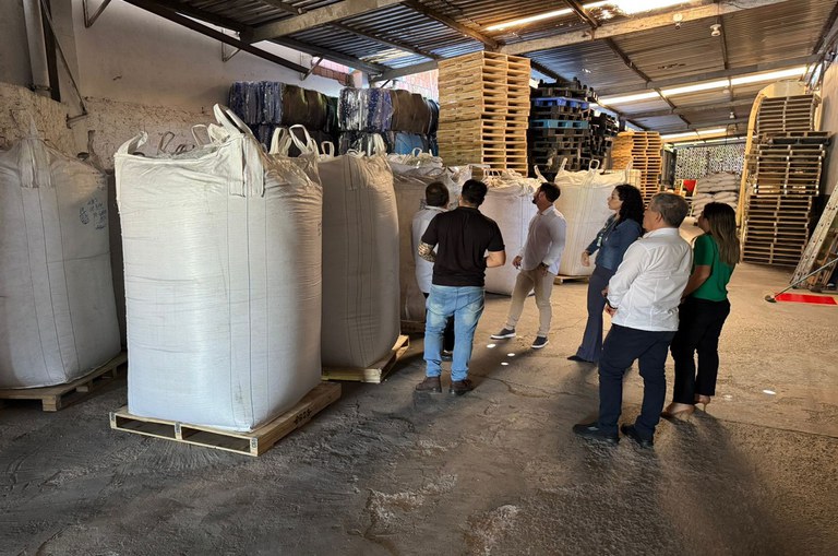 Pessoas em pé no galpão de uma fábrica olhando para sacos grandes com material indefinido