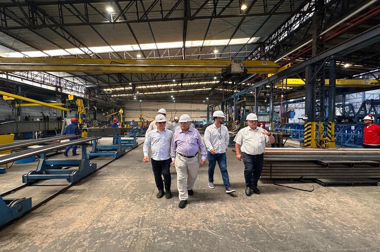 Cinco homens com capacetes de proteção caminham por dentro de uma fábrica de estruturas metálicas. Ao fundo, operários trabalham em máquinas e equipamentos industriais.