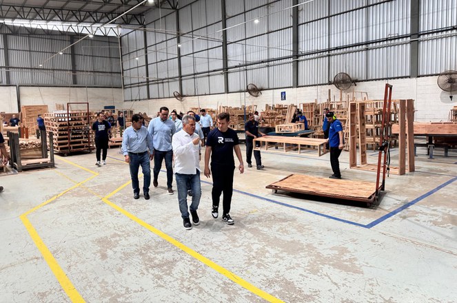 Foto mostra dois homes conversando com um grupo de pessoas atrás e móveis de madeira em produção