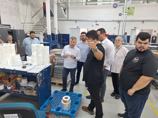 Foto mostra homem segurando um produto adesivo, observado por grupo de pessoar