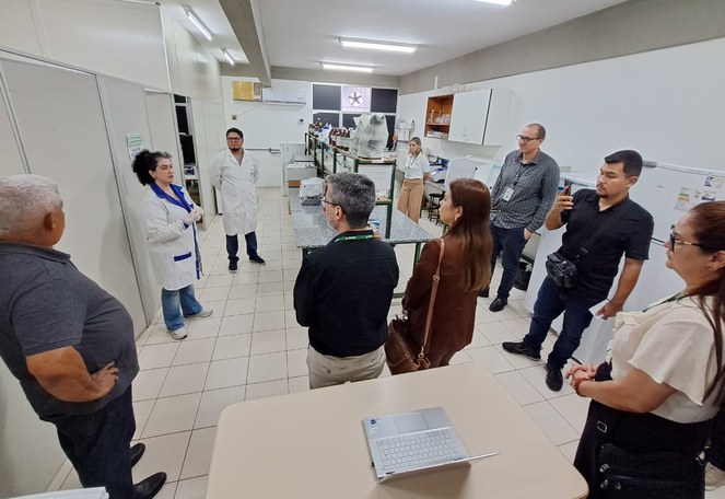Foto mostra mulher de bata branca fazendo uma explanação a visitantes de um laboratório