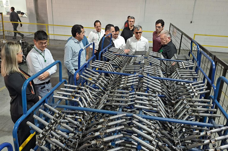 Foto mostra homem de blusa branca e óculos escuros observando, com um grupo de pessoas, umas peças para motocicletas