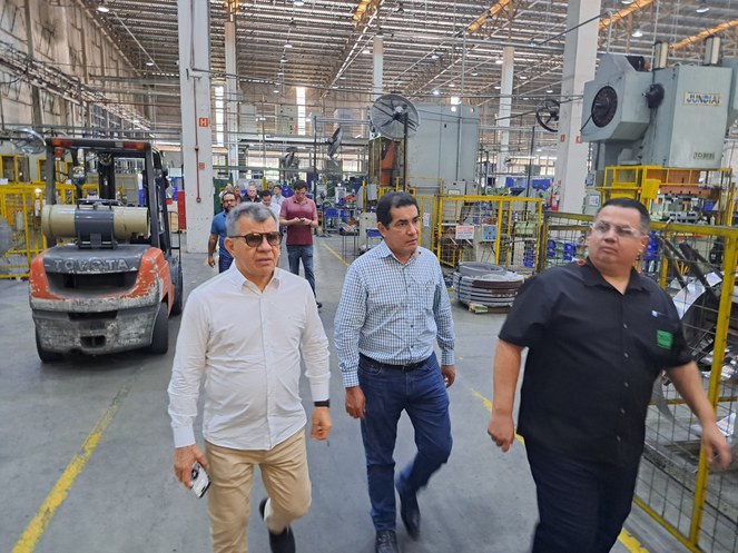 Foto mostra homem de blusa branca e óculos escuros, acompanhado de outros dois homens, em visita a uma fábrica