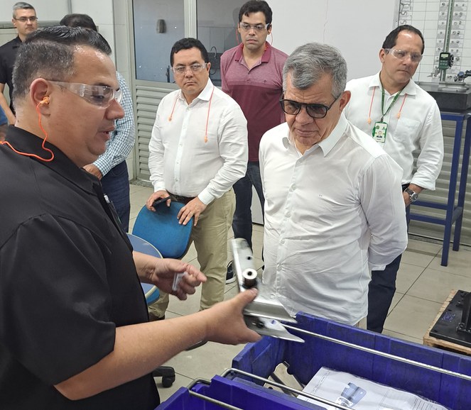 Foto mostra homem de blusa escura e óculos de proteção mostrando peça de motocicleta a outro homem