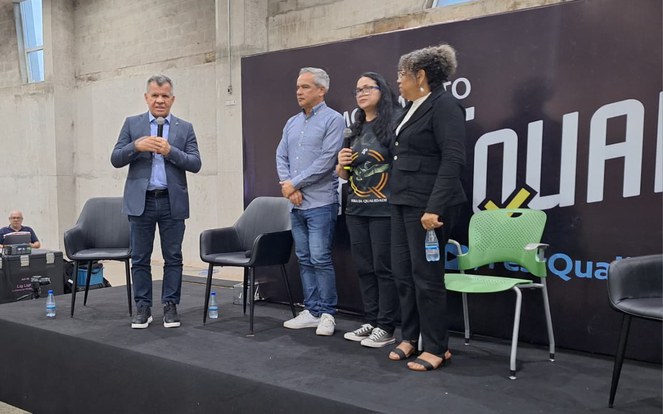 Foto mostra homem com microfone na mão ao lado de três pessoas